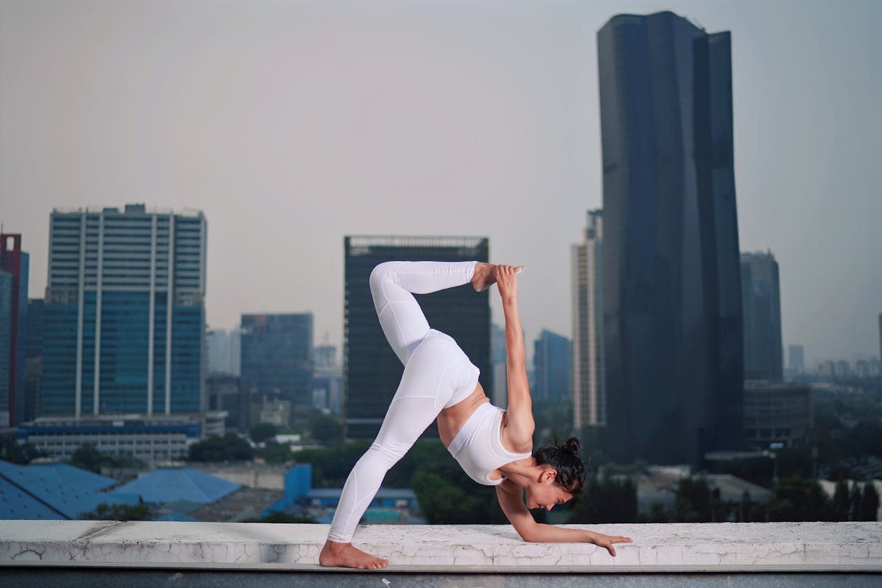 A woman in white activewear performs a yoga pose on a rooftop with an urban skyline backdrop.